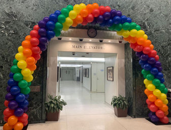 Rainbow balloon arch in pride colors framing the 'Main Elevators' entrance of a marble-walled office lobby with potted plants, corridor and clock overhead.