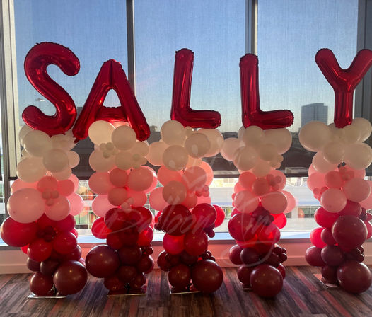 Festive event decor with red foil letter balloons spelling SALLY atop pink, white and red balloon columns by large windows with a downtown skyline view