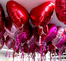 Shiny red and pink heart-shaped foil helium balloons clustered at an indoor ceiling with curly ribbon tails — festive Valentine’s Day party decorations