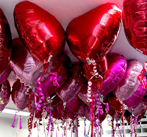 Shiny red and pink heart-shaped foil helium balloons clustered at an indoor ceiling with curly ribbon tails — festive Valentine’s Day party decorations