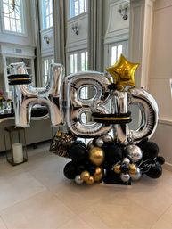 Large indoor birthday balloon display spelling “HBD” in metallic silver letter balloons with black, gold and silver balloon clusters and a gold star, staged in a bright high‑ceiling living room with tall windows and neutral tile floor.