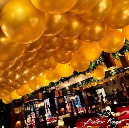 Festive indoor restaurant scene with ceiling packed with shiny gold balloons, holiday evergreen garlands, warm lighting, and cozy red booth seating.