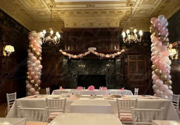 Ornate banquet hall with carved wood fireplace and coffered ceiling decorated for a pink-and-gold celebration, featuring tall balloon columns, floral garland, crystal chandeliers, white chiavari chairs and long banquet tables with blush place settings.