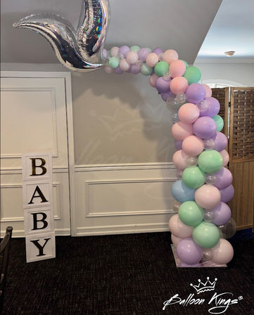 Indoor baby shower balloon arch in pastel pink, lavender and mint with clear accents and a shiny silver mermaid-tail balloon above stacked blocks spelling BABY.