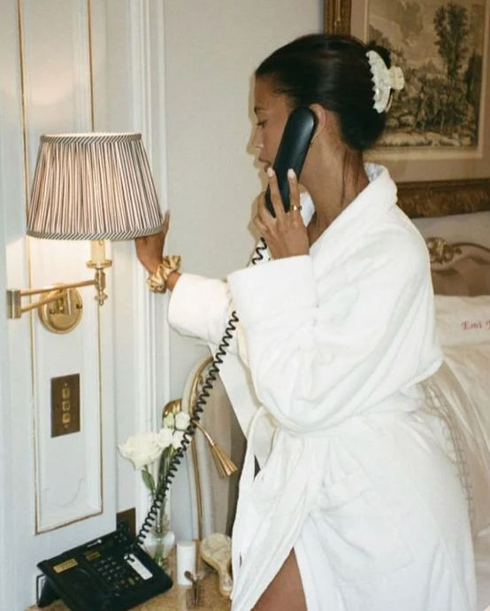 Woman in a white bathrobe speaking on a corded bedside phone, reaching to dim a striped lamp in an elegant, gold-accented hotel room with fresh roses on the nightstand.