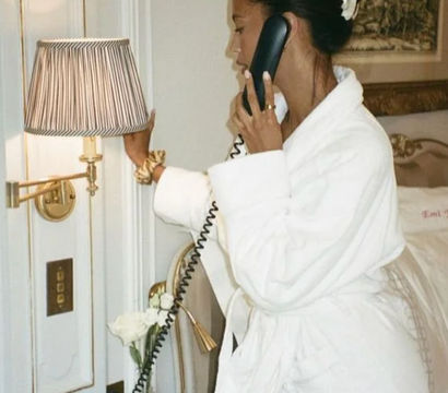 Woman in a white bathrobe speaking on a corded bedside phone, reaching to dim a striped lamp in an elegant, gold-accented hotel room with fresh roses on the nightstand.