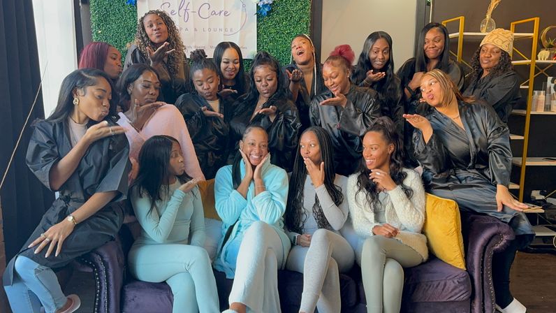 Group of women at a spa self-care party in an indoor lounge, many wearing black robes, gathered on and around a purple sofa; center guest in a light blue robe smiling while others blow kisses under a decorative greenery backdrop.