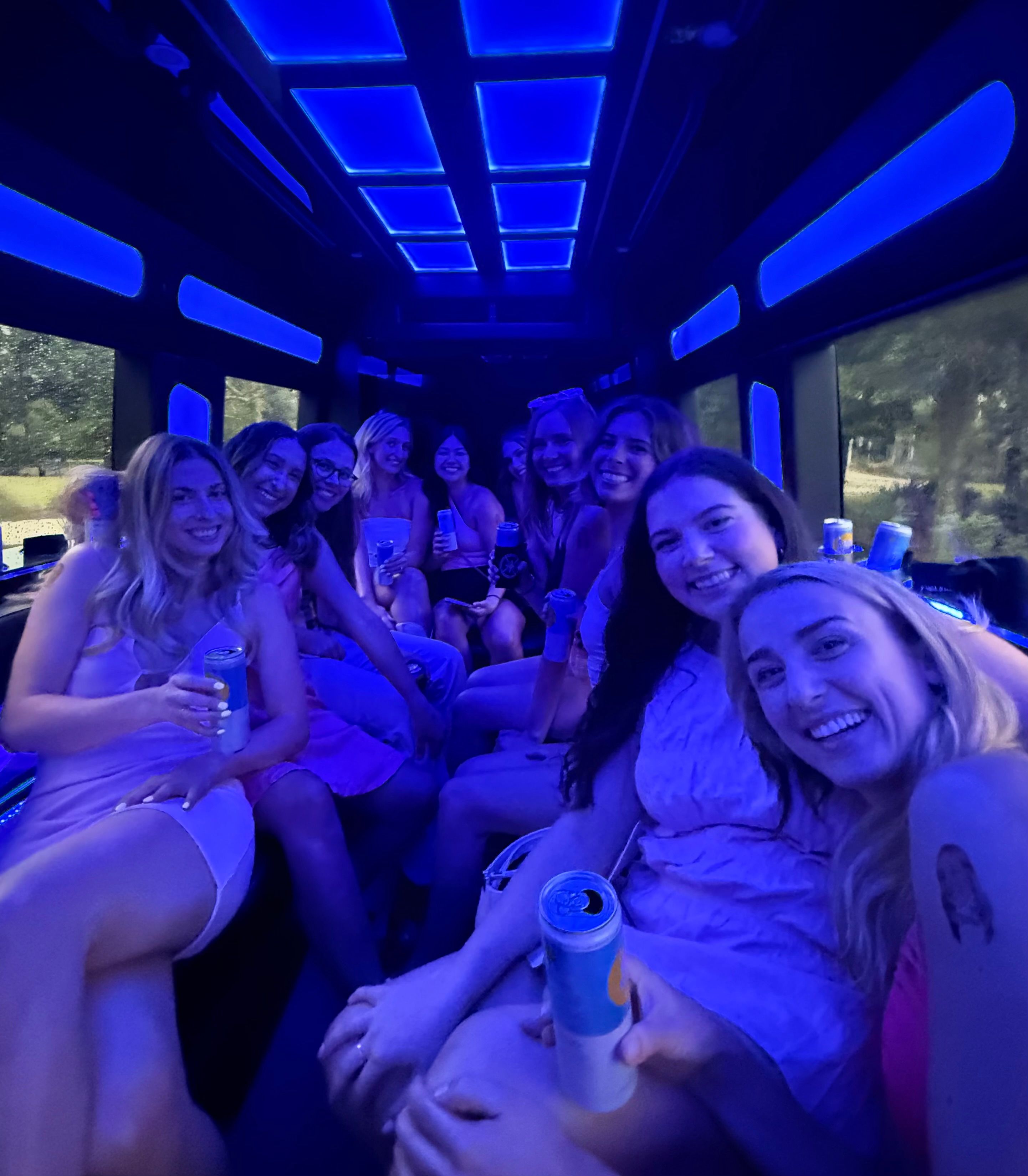 Smiling group of women on a blue LED-lit party bus, holding canned drinks and enjoying a fun night out
