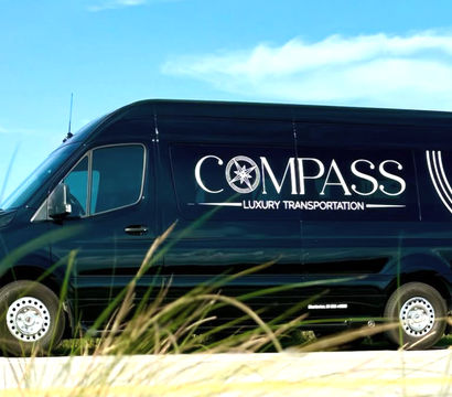 Sleek black luxury shuttle van with a large compass-style logo parked on a sunny coastal road, dune grasses in the foreground and blue sky overhead.