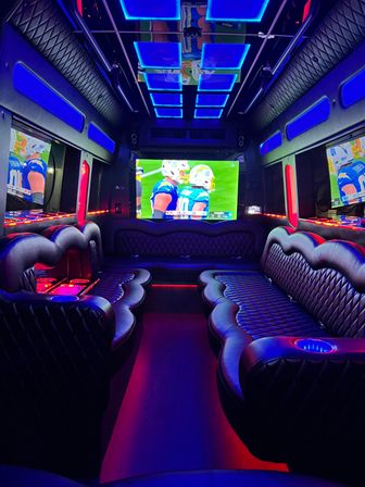 Party bus interior with quilted leather wraparound seating, blue and red LED accent lighting, mirrored ceiling panels and a large flat-screen TV showing a football game — luxury mobile lounge for group entertainment.