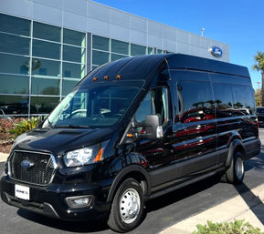 Black Ford high-roof passenger van parked outside a glass-front dealership on a sunny day