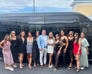 Group photo of one man and several women in summer dresses and casual outfits posing and smiling in front of a sleek black passenger van in a parking lot on a sunny day