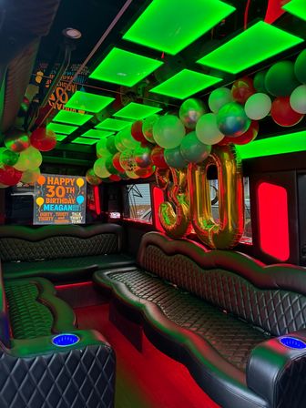Party bus interior with quilted black leather bench seating, neon green LED ceiling panels, red accent lighting, multicolored balloon garland and large gold "30" foil balloons for a birthday celebration.