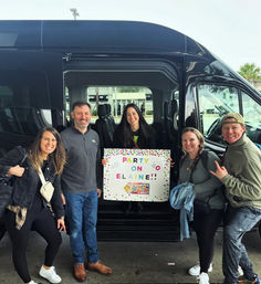 Five smiling adults posing by a black shuttle van at a curb, woman in the center holding a colorful sign that reads “PARTY ON ELAINE!!” — friends ready for a fun group celebration and ride.