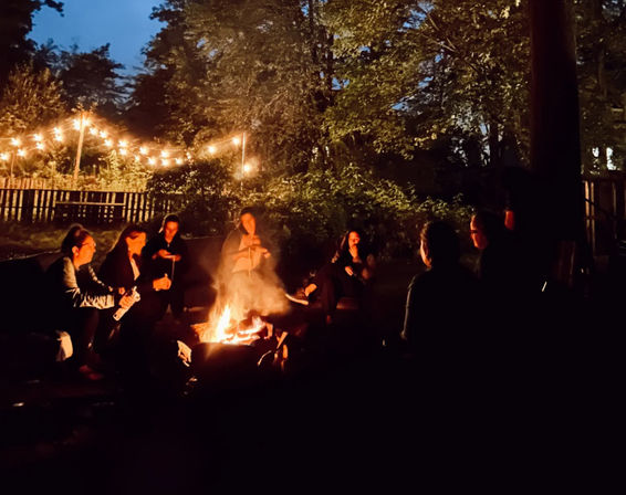 Cozy backyard summer evening: friends gathered around a glowing campfire beneath string lights and tall trees against a twilight sky.