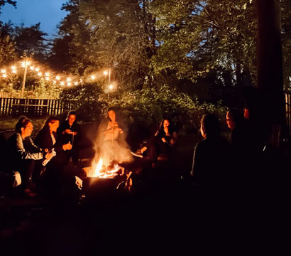 Cozy backyard summer evening: friends gathered around a glowing campfire beneath string lights and tall trees against a twilight sky.