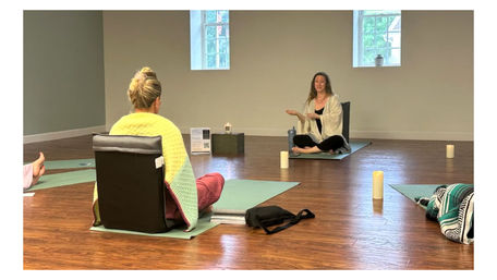 Cozy indoor yoga and meditation class in a bright studio — instructor seated cross-legged guiding a small group on mats and floor chairs with candles.
