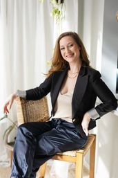 Relaxed lifestyle portrait of a smiling woman in a black blazer and satin navy pants seated on a woven chair in a bright sunlit room