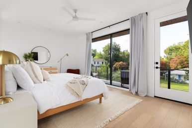 Bright modern master bedroom with white linens on a wood platform bed, gold bedside lamp, round wall mirror and floor-to-ceiling sliding glass doors opening to a balcony with lush suburban backyard views.