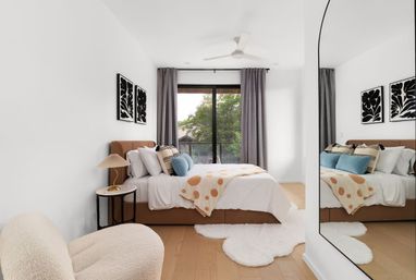 Sunlit modern bedroom with tan upholstered bed, polka-dot throw and blue accent pillows, sliding glass door to a leafy balcony, fluffy rugs and large wall mirror.