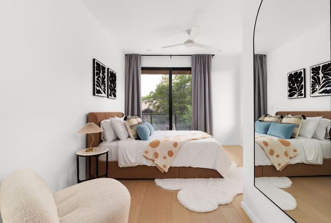 Sunlit modern bedroom with tan upholstered bed, polka-dot throw and blue accent pillows, sliding glass door to a leafy balcony, fluffy rugs and large wall mirror.