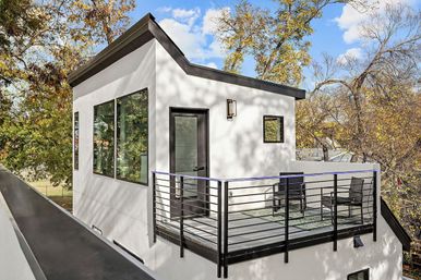 Chic white rooftop studio with sloped roof, large windows and glass door opening to a black metal-railed balcony with chairs, surrounded by autumn trees under a blue sky.