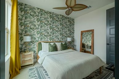 Sunlit modern tropical-inspired bedroom with botanical leaf wallpaper, green velvet headboard, wooden ceiling fan, mustard curtain, matching bedside tables, floor mirror and patterned rug.