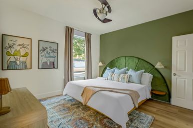 Bright modern bedroom with green velvet fan-shaped headboard, king bed with white linens and decorative pillows, patterned area rug, wood floors, framed wall art and large window letting in natural light.