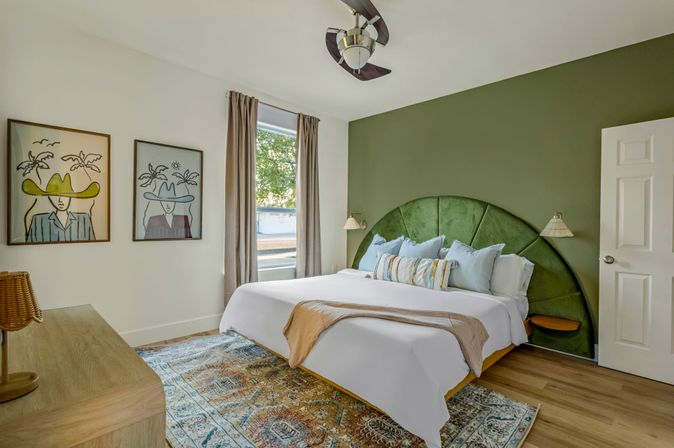 Bright modern bedroom with green velvet fan-shaped headboard, king bed with white linens and decorative pillows, patterned area rug, wood floors, framed wall art and large window letting in natural light.