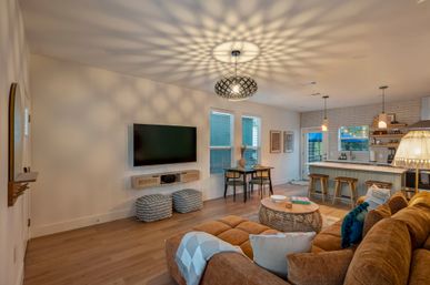 Open-concept modern living room and kitchen with a brown sectional sofa, round rattan coffee table, wall-mounted TV, patterned pendant light casting starburst shadows on the ceiling, breakfast bar with wooden stools, dining nook by windows, and light wood floors.