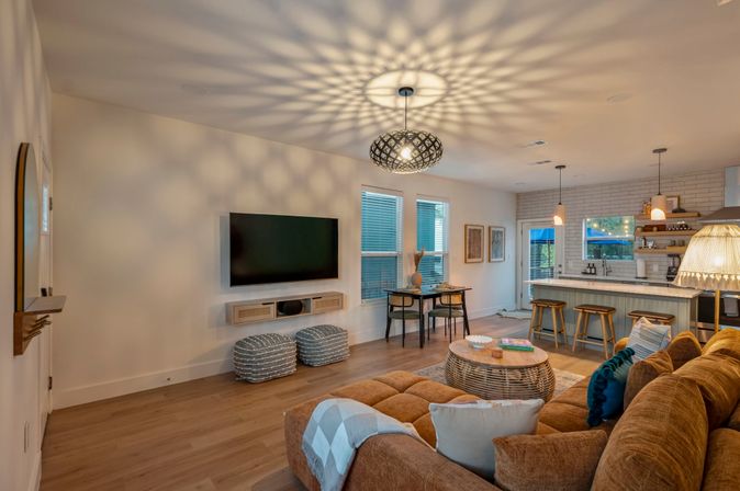 Open-concept modern living room and kitchen with a brown sectional sofa, round rattan coffee table, wall-mounted TV, patterned pendant light casting starburst shadows on the ceiling, breakfast bar with wooden stools, dining nook by windows, and light wood floors.