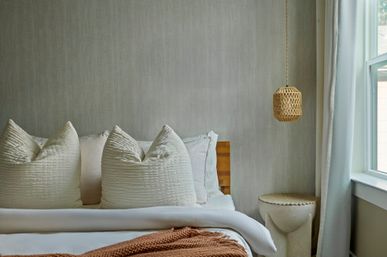Cozy neutral bedroom interior with layered white bedding, three plump textured pillows, a woven hanging pendant, round stone bedside stool and sunlit window
