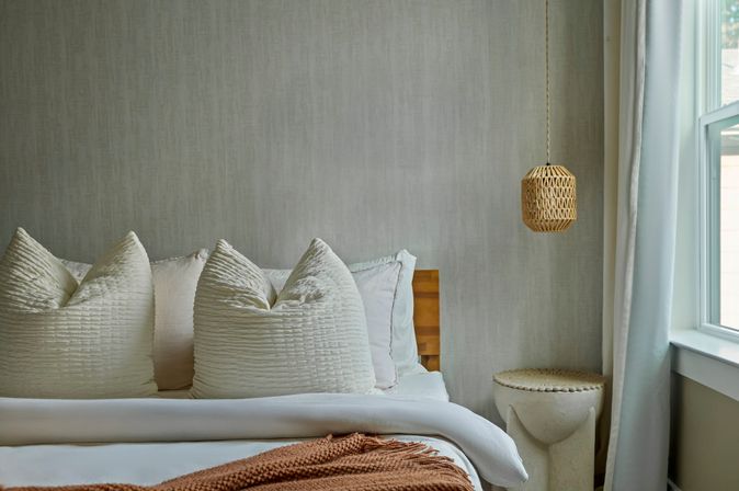 Cozy neutral bedroom interior with layered white bedding, three plump textured pillows, a woven hanging pendant, round stone bedside stool and sunlit window