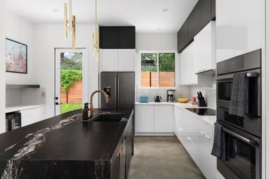 Modern open-concept kitchen with a black marble island and gold faucet, glossy white cabinets, stainless refrigerator and double oven, pendant lights, concrete floor, and a door and window framing a fenced backyard.