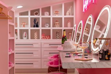 Pink glam dressing room with illuminated oval vanity mirrors, marble countertop holding champagne bottles and glasses, a white 'bride' hat, pink stools and cubby shelves of decorative objects