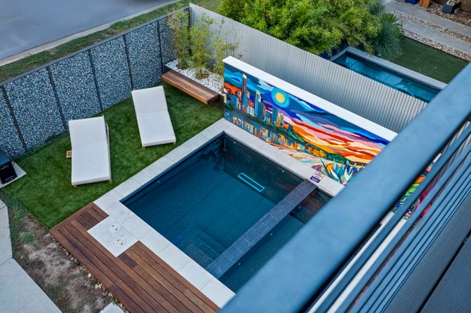 Aerial view of a modern backyard plunge pool with a colorful cityscape mural, two white lounge chairs on artificial turf, and wooden decking.