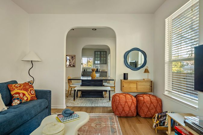 Sunlit modern-boho living room with blue sofa and colorful pillows, wood floors, orange woven poufs and round blue mirror; arched doorway opens to a dining area with bench seating and kitchen beyond.