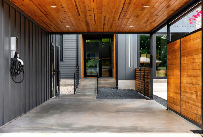 Modern residential carport and covered entry with warm wood ceiling, concrete floor, wall-mounted EV charger, sleek metal siding, and wooden privacy panels leading to a glass front door.