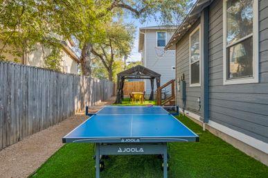 Blue outdoor ping-pong table on artificial turf in a narrow residential side yard, flanked by a wooden fence and gray house siding, with a gazebo seating area and string lights overhead.