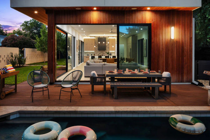 Modern wood-paneled house with sliding glass doors opening to a lit living room, poolside wooden deck with outdoor dining table, lounge chairs and colorful inflatable rings in the pool at dusk.