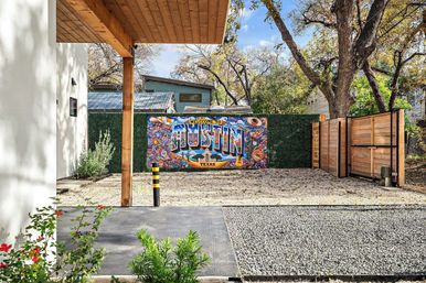 Vibrant “Welcome to Austin, Texas” mural on a green hedge in a modern fenced backyard with gravel driveway, wooden gate, patio and trees