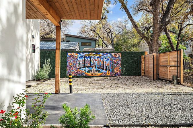 Vibrant “Welcome to Austin, Texas” mural on a green hedge in a modern fenced backyard with gravel driveway, wooden gate, patio and trees