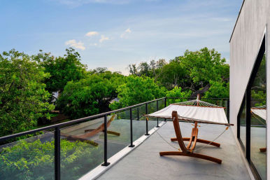 Modern rooftop balcony with a hammock on a wooden stand and glass railing overlooking a lush treetop canopy under a blue sky
