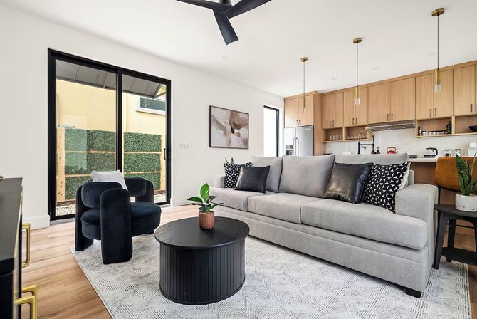 Bright modern open-plan living room and kitchen — gray sofa with patterned pillows, black accent chair, round black coffee table with potted plant, wood cabinets, pendant lights, and sliding glass door to a small patio.