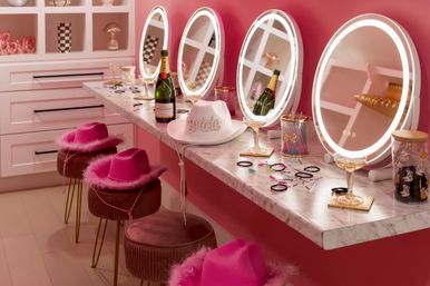 Pink glam vanity makeup station with four illuminated round mirrors, marble counter holding champagne bottles and coupe glasses, feathered hot-pink hats on velvet stools, and scattered hair ties and beauty accessories — playful bachelorette/bridal dressing area.