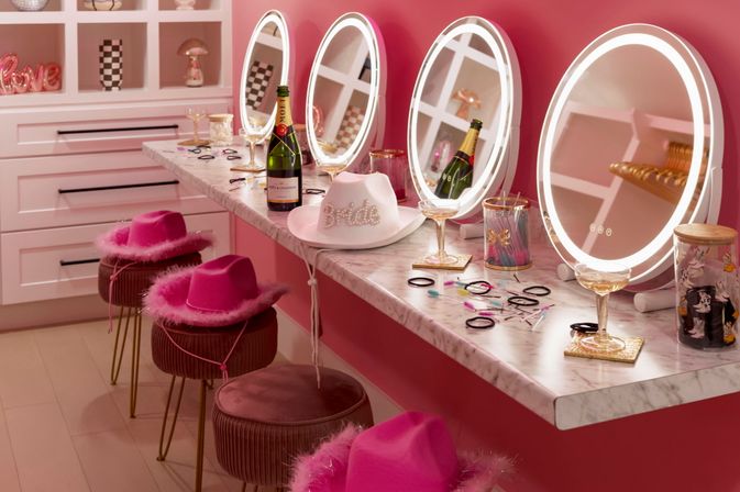 Pink glam vanity makeup station with four illuminated round mirrors, marble counter holding champagne bottles and coupe glasses, feathered hot-pink hats on velvet stools, and scattered hair ties and beauty accessories — playful bachelorette/bridal dressing area.