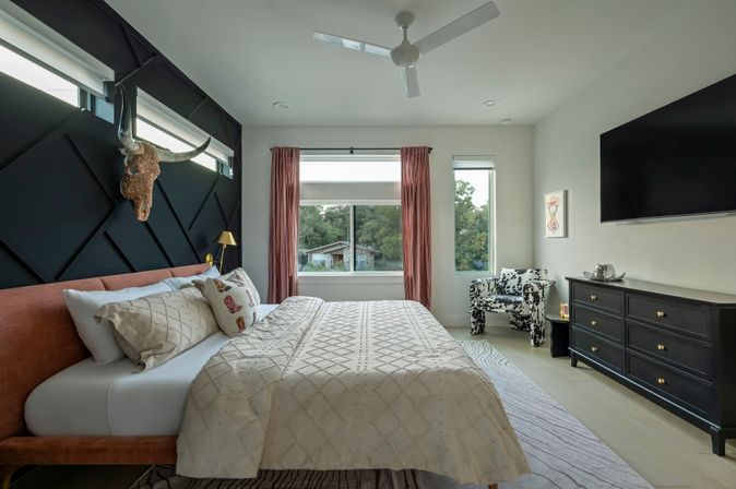 Stylish contemporary bedroom with black geometric accent wall and mounted longhorn skull, coral upholstered bed with patterned bedding, ceiling fan, pink curtains framing a window with leafy suburban view, cow-print armchair and black dresser with wall-mounted TV.