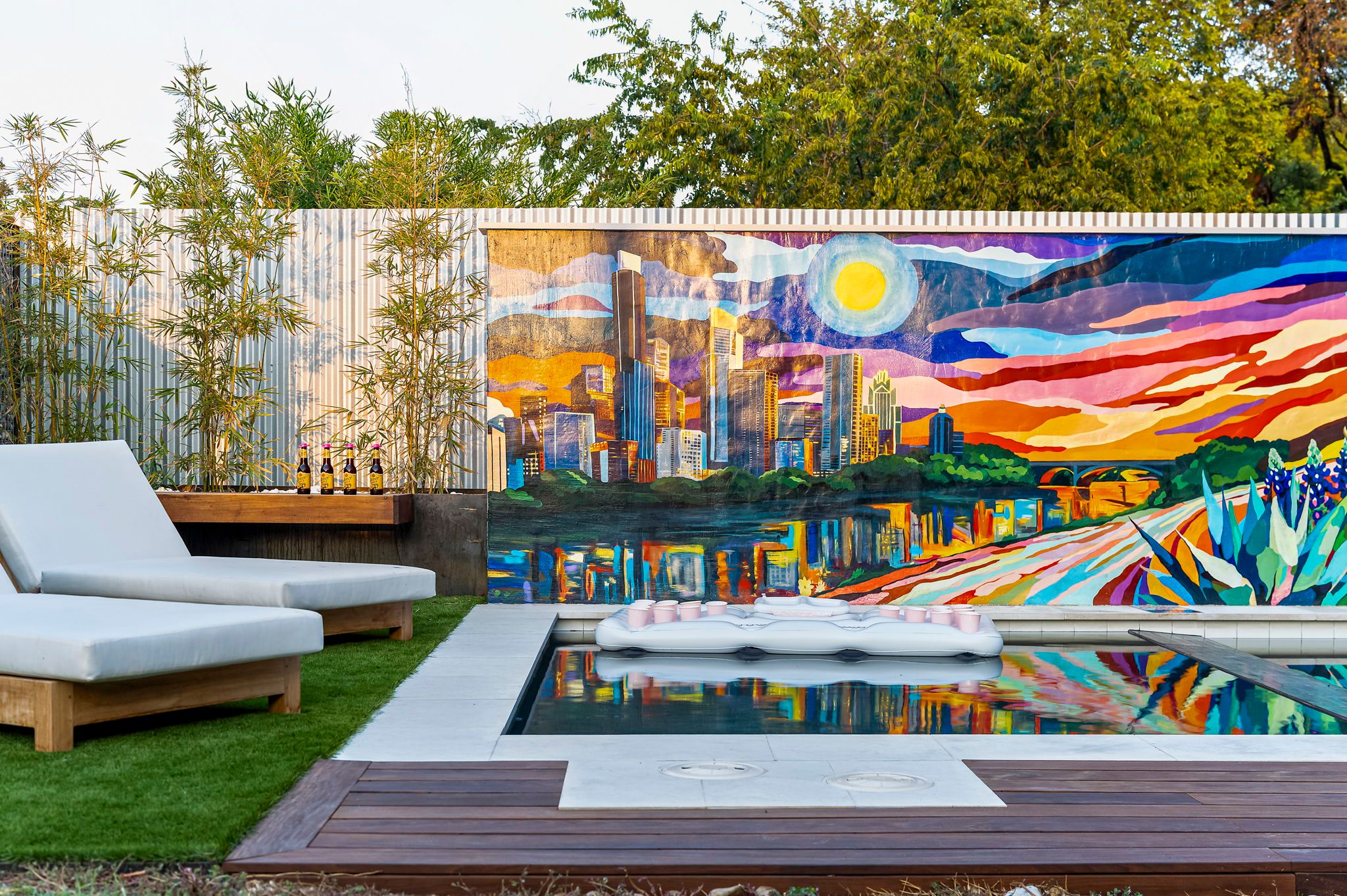 Urban backyard pool with white cushioned lounge chairs, bamboo plants and wooden deck, colorful sunset city skyline mural reflecting in the water and a white inflatable pool float.