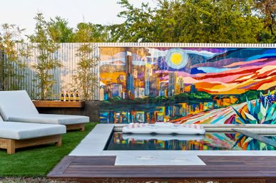 Urban backyard pool with white cushioned lounge chairs, bamboo plants and wooden deck, colorful sunset city skyline mural reflecting in the water and a white inflatable pool float.