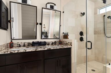 Modern spa-like bathroom with double vanity, granite countertop, dark wood cabinets, black fixtures and glass-enclosed beige tile shower.